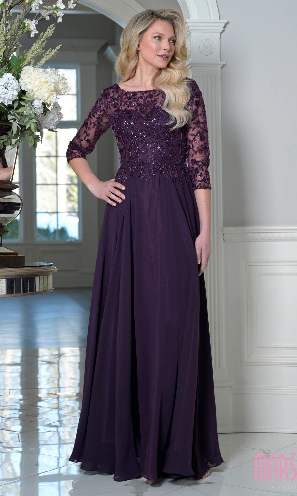 A woman stands indoors near a large window and floral arrangement, wearing the Marsoni M340SL long formal dress in eggplant with a modest beaded-embroidered bodice and A-line chiffon skirt.